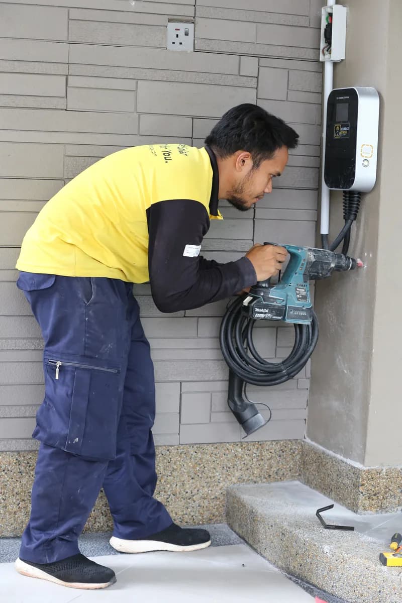 An installer in a hard hat drilling into an exterior wall beside a newly mounted charger